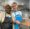 two women smiling one in a black apron one in a blue apron holding a pan in a kitchen